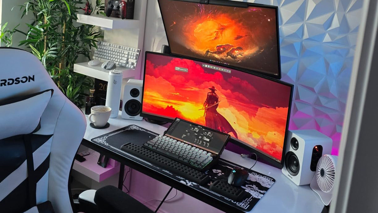 Modern gaming setup with dual monitors displaying vibrant red and orange artwork, mechanical keyboard with large desk mat, white gaming chair, desktop speakers, and RGB lighting in a clean workspace